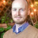 Dr. Brian S. Sedgeley, PsyD, a clinical psychologist and founder of Bay Psychology Group, Inc., wearing a brown sweater and checkered shirt, smiling warmly in a garden setting with soft lighting in the background.