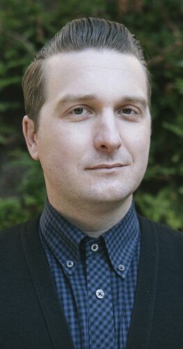 Portrait of Ryan LaPlant, AMFT with short, slicked-back hair, wearing a checkered shirt and a dark sweater, standing in front of a blurred, green leafy background.