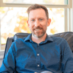 Professional headshot of Nicholas Hack, PsyD, seated in a chair, smiling warmly in a casual blue patterned shirt, with a soft natural light from the window behind him.