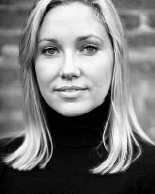 Black and white portrait of Jenny Werba, LMFT, APCC, a licensed marriage and family therapist and associate professional clinical counselor. Jenny has a calm expression and is wearing a black turtleneck, standing against a textured brick wall. width=