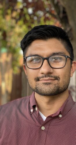 A professional headshot of Dr. Sahil Sharma, PsyD, standing outdoors. He is wearing glasses and a maroon button-up shirt with a calm and approachable expression. The background features soft, blurred greenery and trees, adding a natural setting to the image.