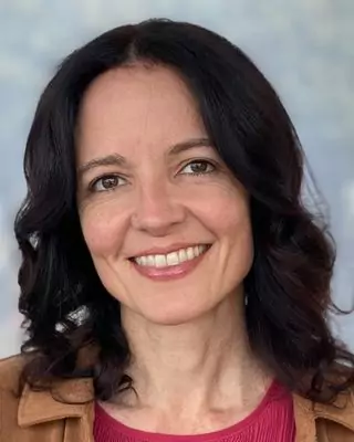 A professional headshot of Anna Guha, LMFT, smiling warmly. She is wearing a red top with a light brown jacket, with a soft background of muted blue and gray tones. width=