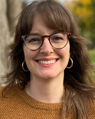 A professional headshot of Danielle Gotwalt, AMFT, smiling warmly at the camera. She is wearing glasses, hoop earrings, and a brown sweater, with an outdoor background blurred behind her. width=