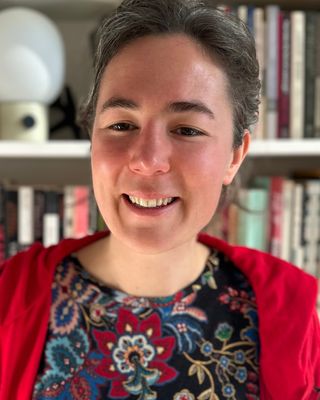 A professional indoor headshot of Katherine Lund, LMFT, smiling gently. She is wearing a colorful floral-patterned top with a red cardigan, standing in front of a bookshelf filled with books. width=