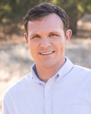 A professional outdoor headshot of Thomas Darksmith, LMFT, smiling warmly at the camera. He is wearing a light blue button-up shirt, with a natural background of trees and sunlight blurred behind him. width=