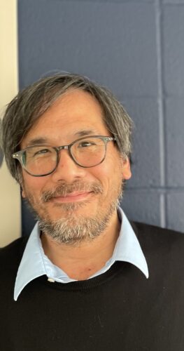 Michael Cheng, MA, a psychological associate at Bay Psychology Group, Inc., stands smiling in front of a navy-blue wall. He has salt-and-pepper hair, a short beard, and round glasses, and wears a light-blue collared shirt under a black sweater.